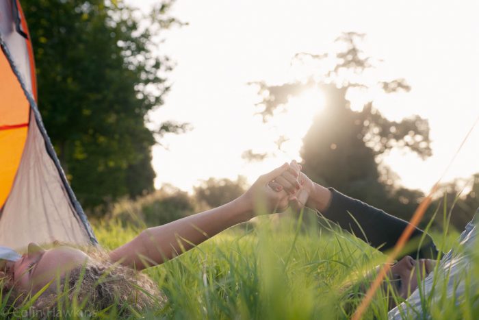Lifestyle photography by Colin Hawkins of two young female friends who are camping and holding hands as the sun sets whilst laying down in a field