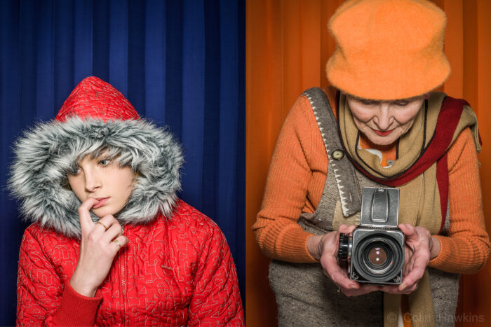 Advertising portrait photography of two women of different ages by Colin Hawkins- young girl with finger in mouth against blue background and senior woman wearing orange clothes against an orange background taking a photograph with an old Hasselblad camera.
